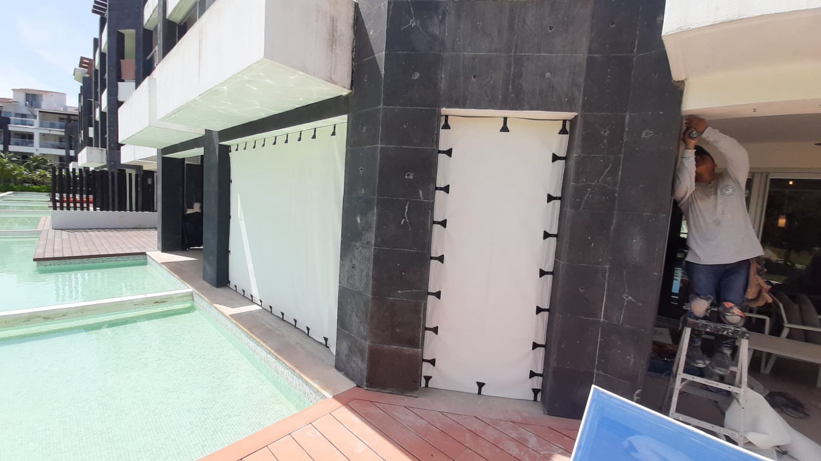 Installer on a ladder securing a fabric screen with black clips beside a residential swimming pool.