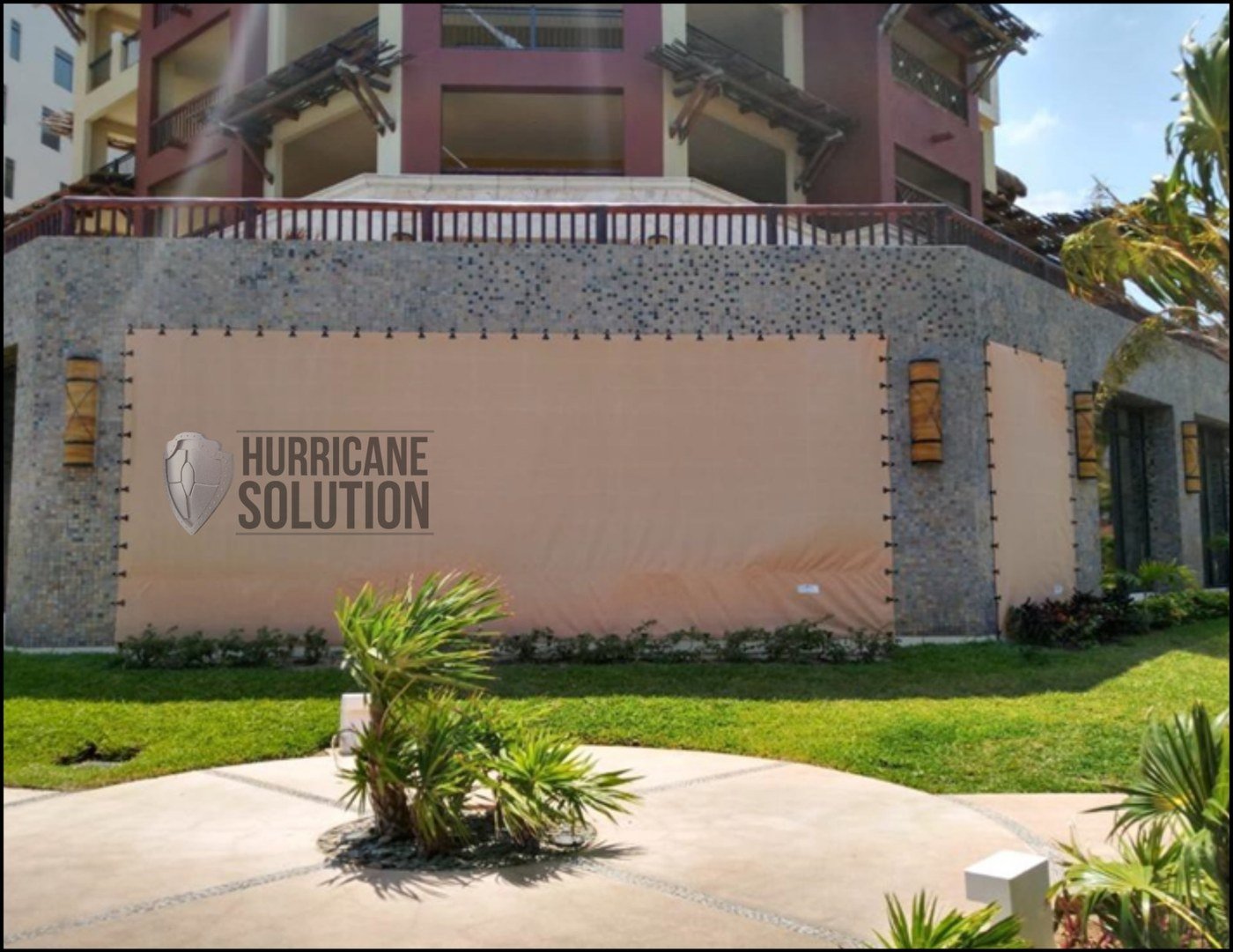 Hurricane Solution screen covering a wide opening below a balcony, surrounded by stone walls and landscaping.